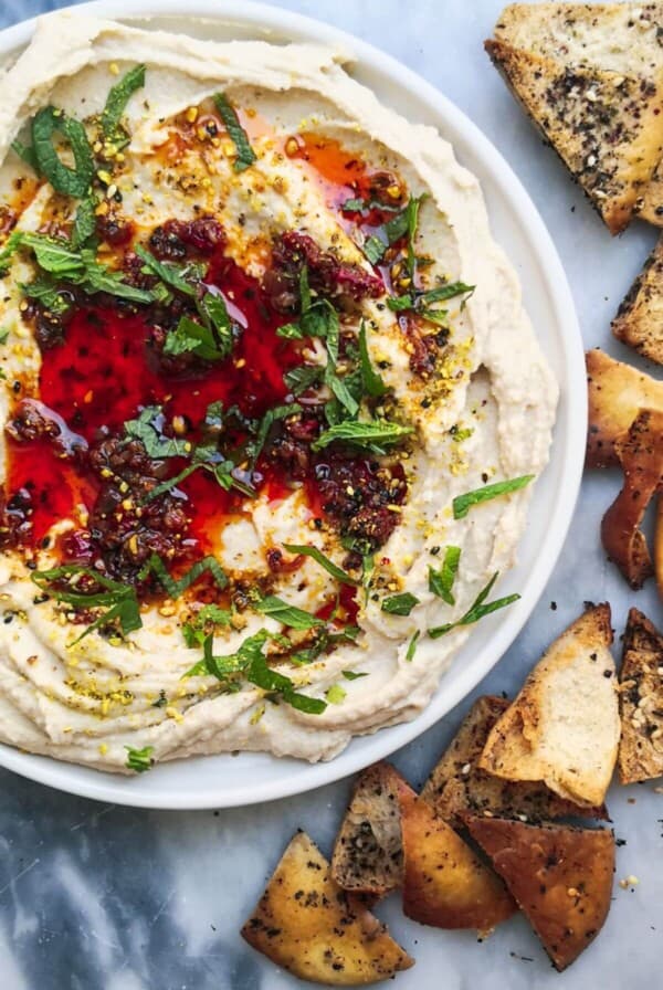 A small white plate of chilli oil topped super smooth hummus, with pita chips on the side.