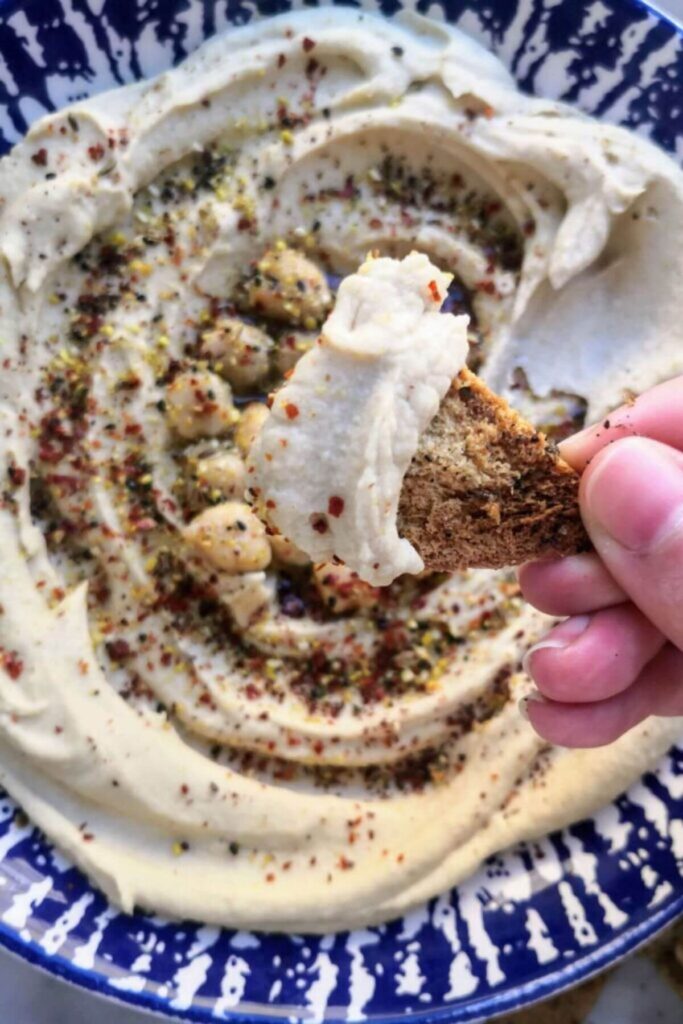 Close up of creamy hummus on a pita crisp with bowl of hummus in the background.