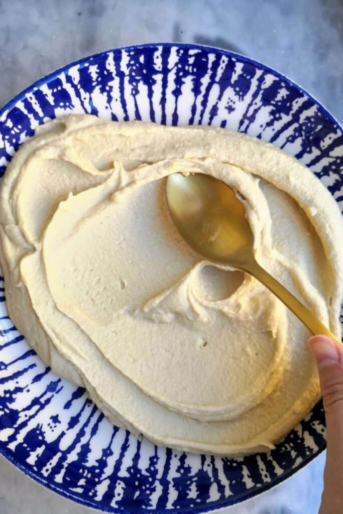 Creamy oil free hummus being smeared onto a blue and white striped plate with a gold spoonn