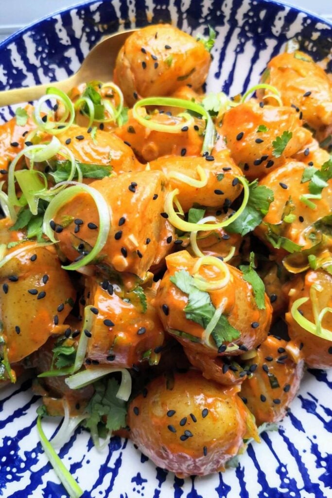 Orange gochujang potatoes in a white and blue bowl with spring onions and black sesame seeds.