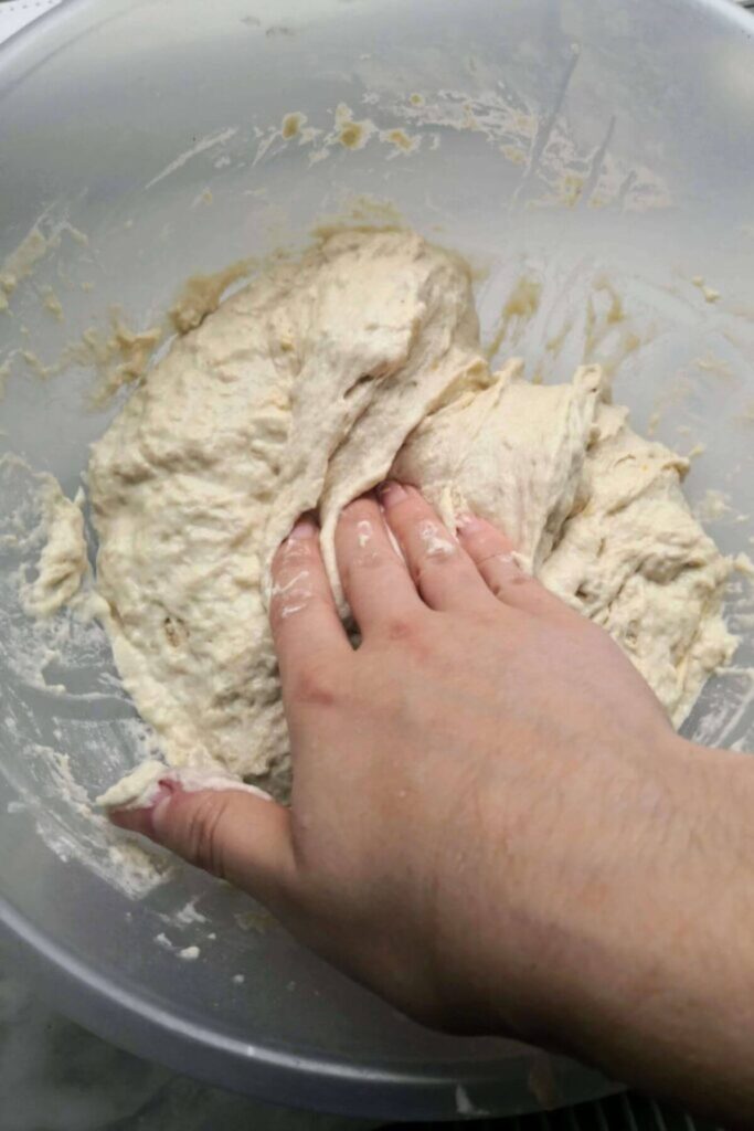 Hand pressing down on the middle of focaccia dough in a large clear mixing bowl.