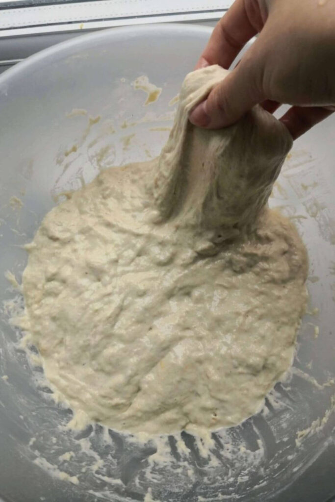 Hand pulling up a portion of focaccia dough in a large, clear mixing bowl.