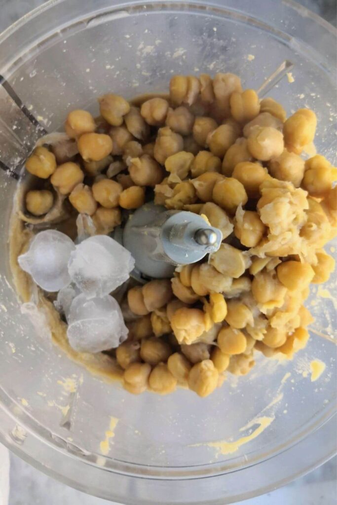 Chickpeas, ice cubes and whipped tahini in the food processor bowl