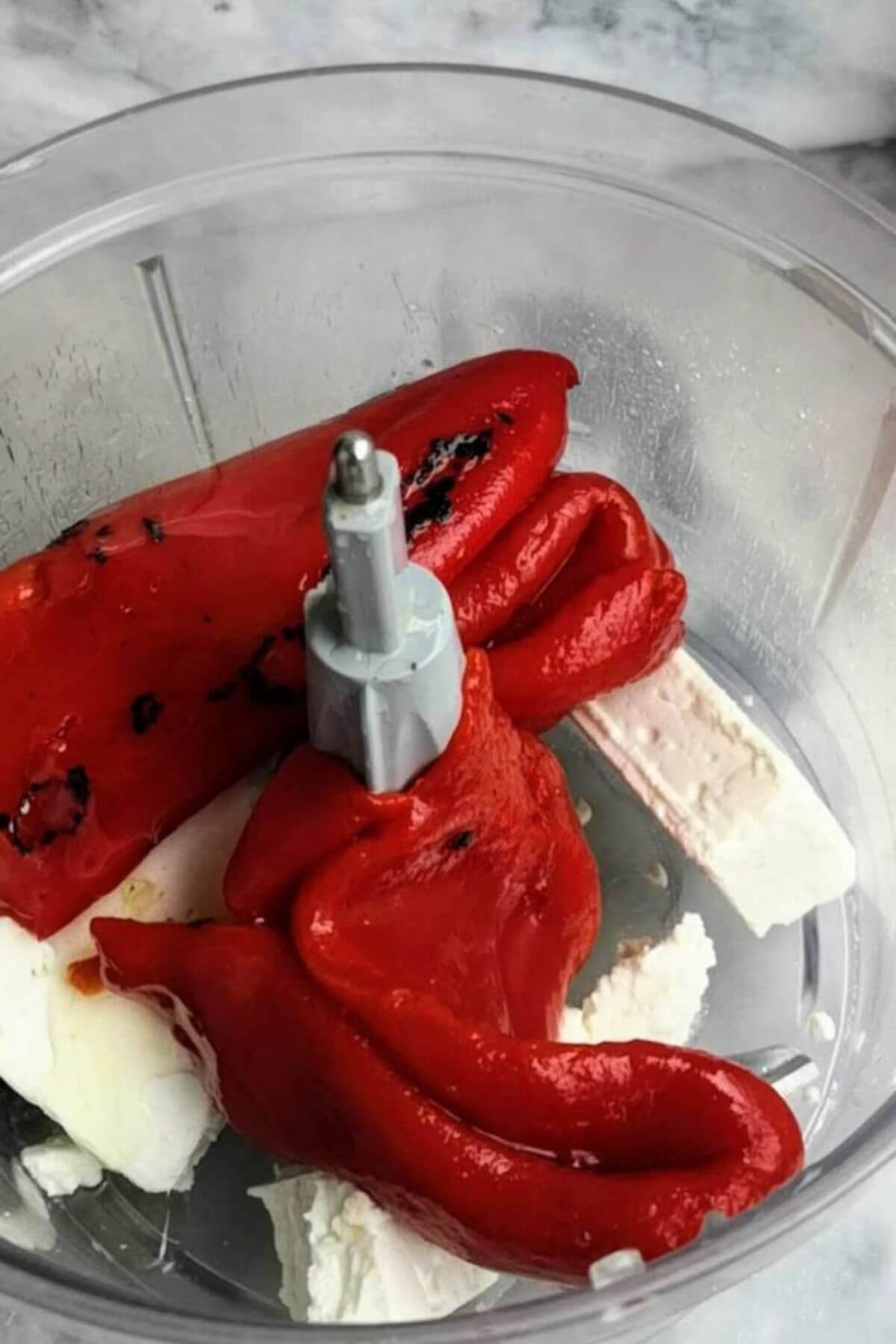 Red pepper and feta pieces in the bowl of a food processor with clear sides