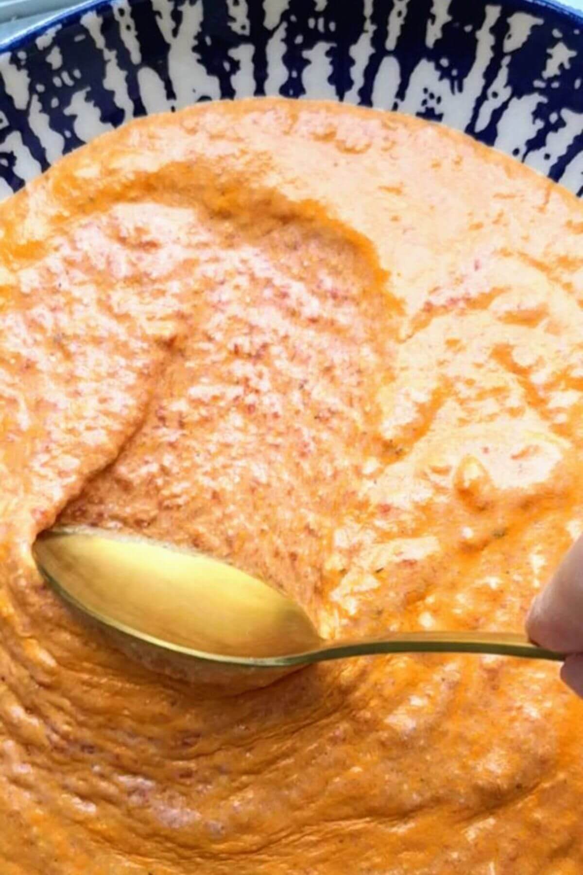 Small gold spoon smoothing red pepper feta dip in a white and blue bowl.