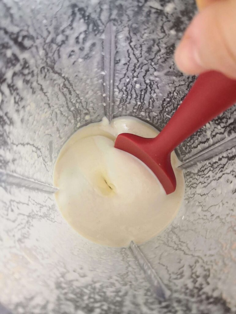 A red spoon about to give the vegan aioli a stir in a clear sided blender looking down from above.