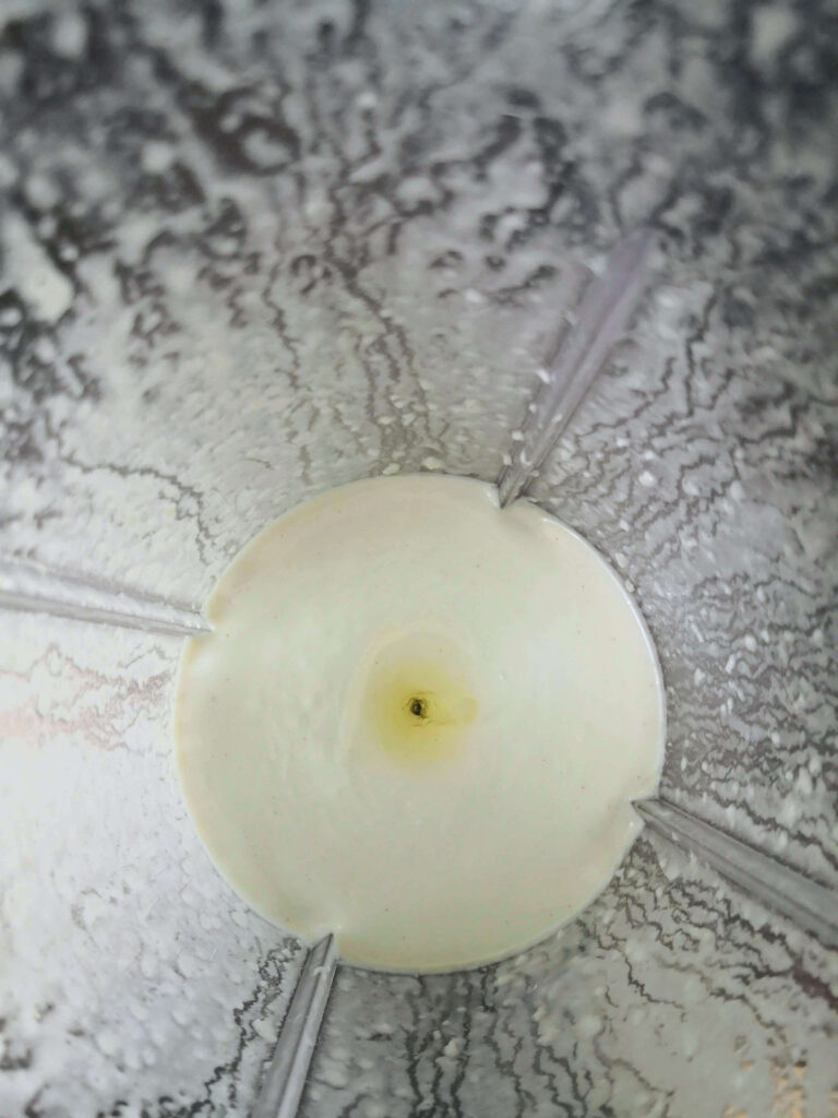 White vegan garlic aioli in a clear sided blender, with a pool of oil collecting on the top in the middle, looking down from above.