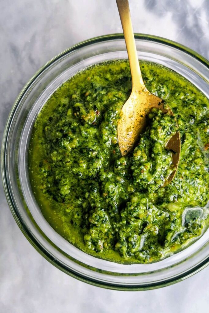 Homemade basil pesto in a small glass bowl, with a gold spoon on a grey marble background.