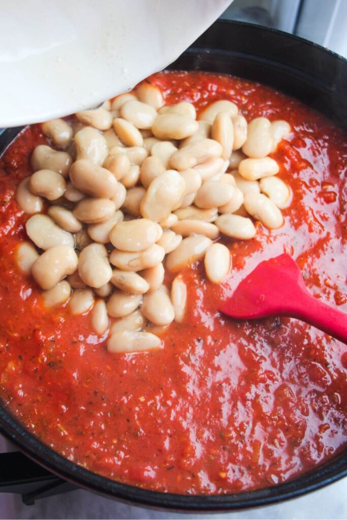 Butter beans being poured into tomato sauce in a large black pan.