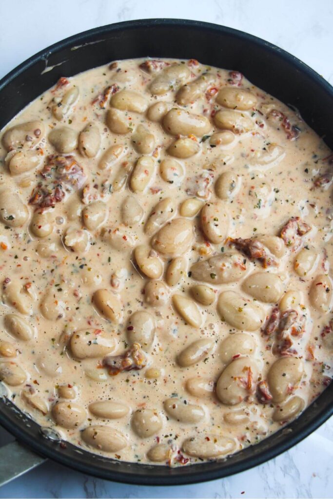 White beans stirred into creamy sundried tomato sauce in a black pan.