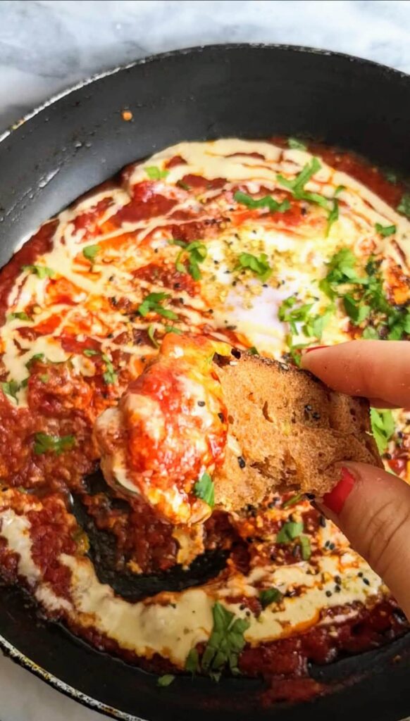 Hand holding up a piece of bread covered in shakshuka sauce.