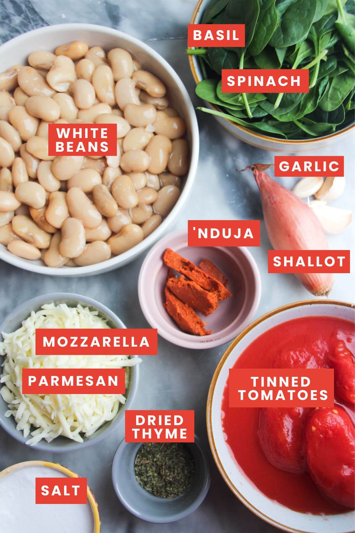Ingredients for cheesy white bean tomato bake laid out on a grey marble background and labelled.