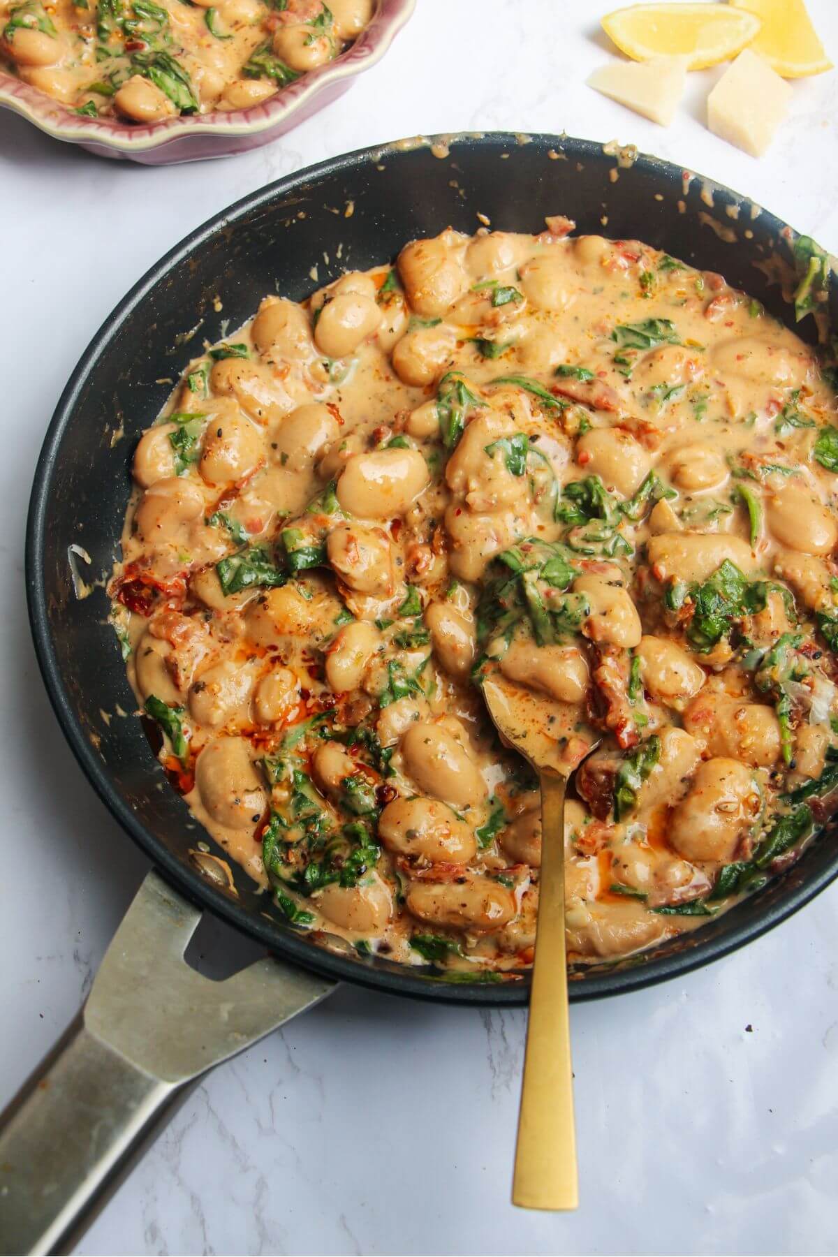 Gold spoon in a pan with creamy sundried tomato white beans, with a small dish of white beans in the background.