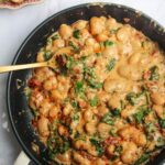 Gold spoon in a pan with creamy sundried tomato white beans, with a small dish of white beans in the background.