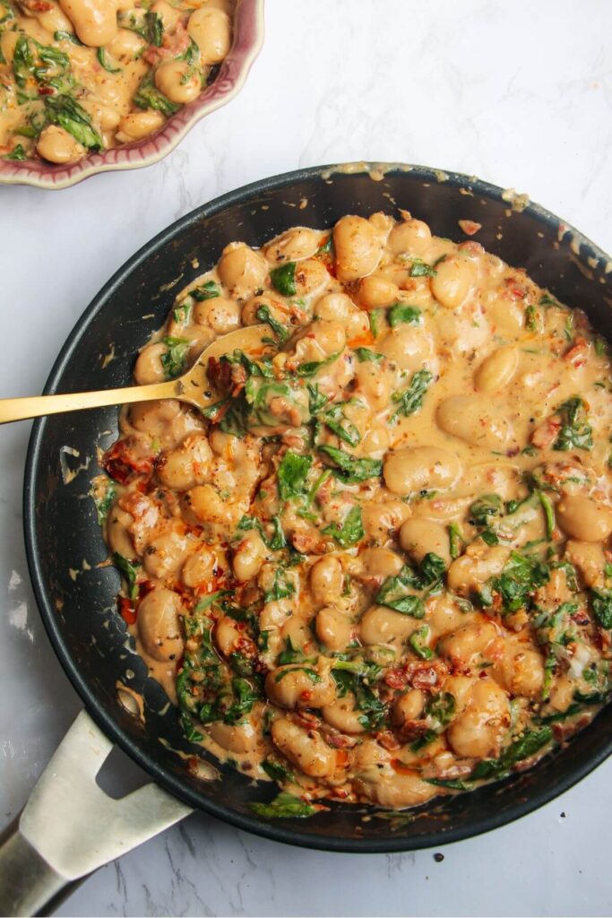 Gold spoon in a pan with creamy sundried tomato white beans, with a small dish of white beans in the background.