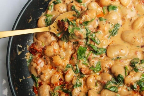 Gold spoon in a pan with creamy sundried tomato white beans, with a small dish of white beans in the background.