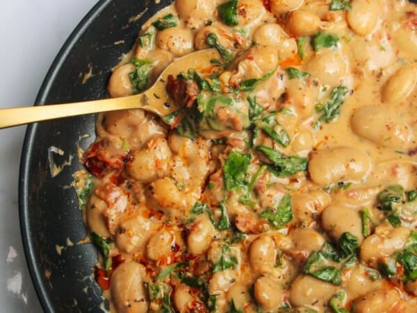 Gold spoon in a pan with creamy sundried tomato white beans, with a small dish of white beans in the background.