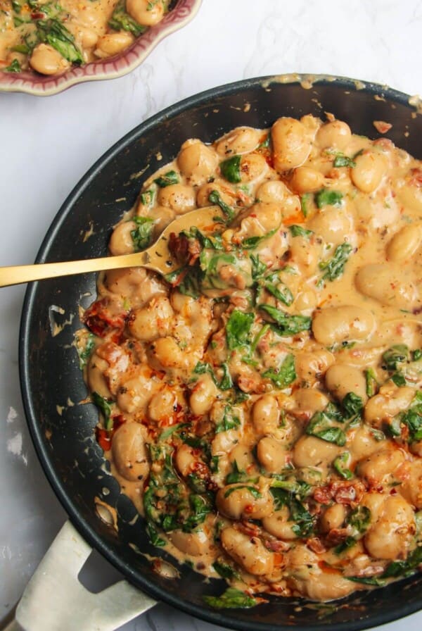 Gold spoon in a pan with creamy sundried tomato white beans, with a small dish of white beans in the background.