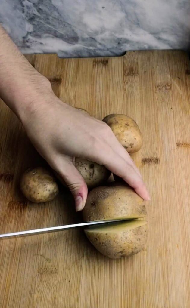 Potato being cut into chunks with large knife on a wooden board.