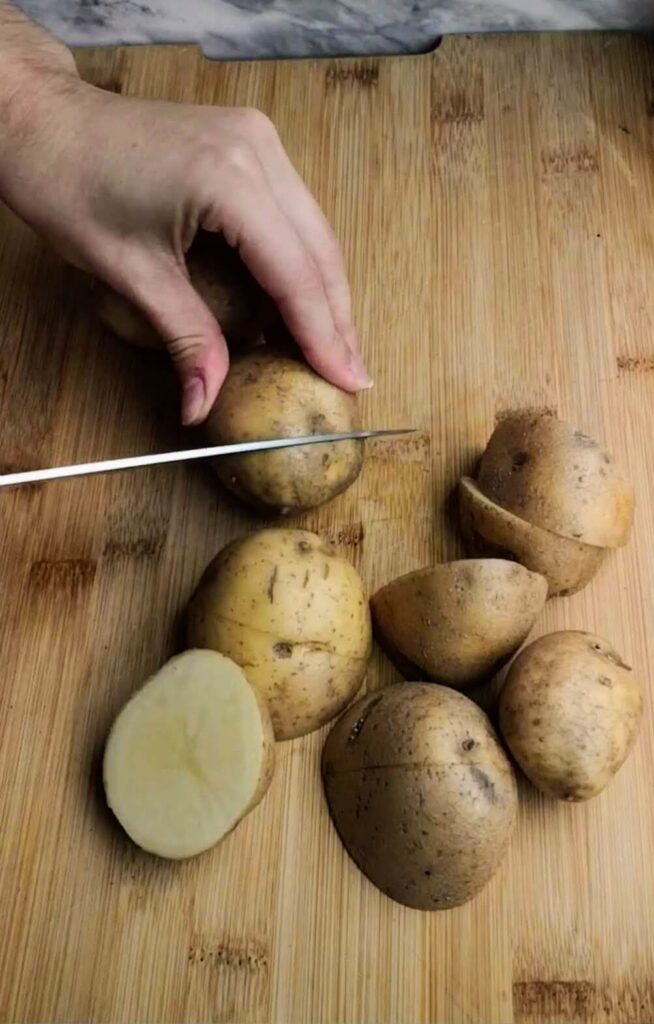 Small potato being cut into chunks on a wooden board with six other potatoes on board.