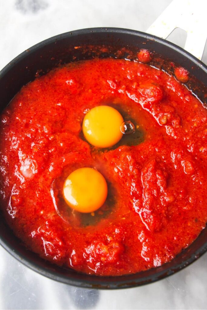 Two eggs cracked into tomato sauce in a small black pan.