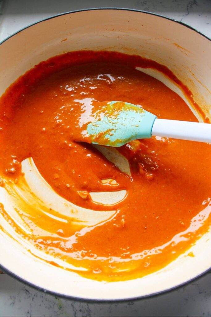 Blue and white spatula stirring bright orange gochujang sauce in a large white skillet.