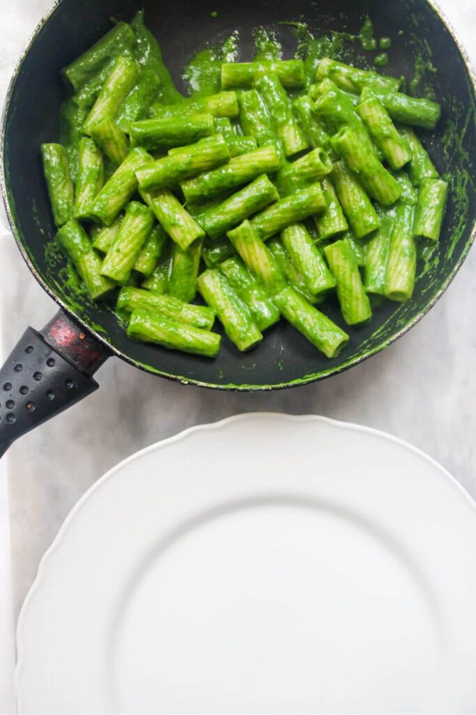 Green pasta in a small pan with a white plate in front of it, ready to be served.