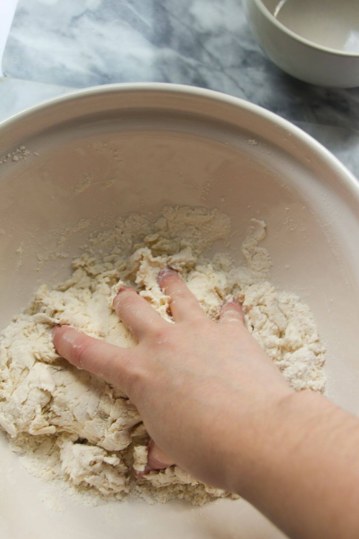 A hand squeezing flour and yogurt dough together in a large ceramic bowl.