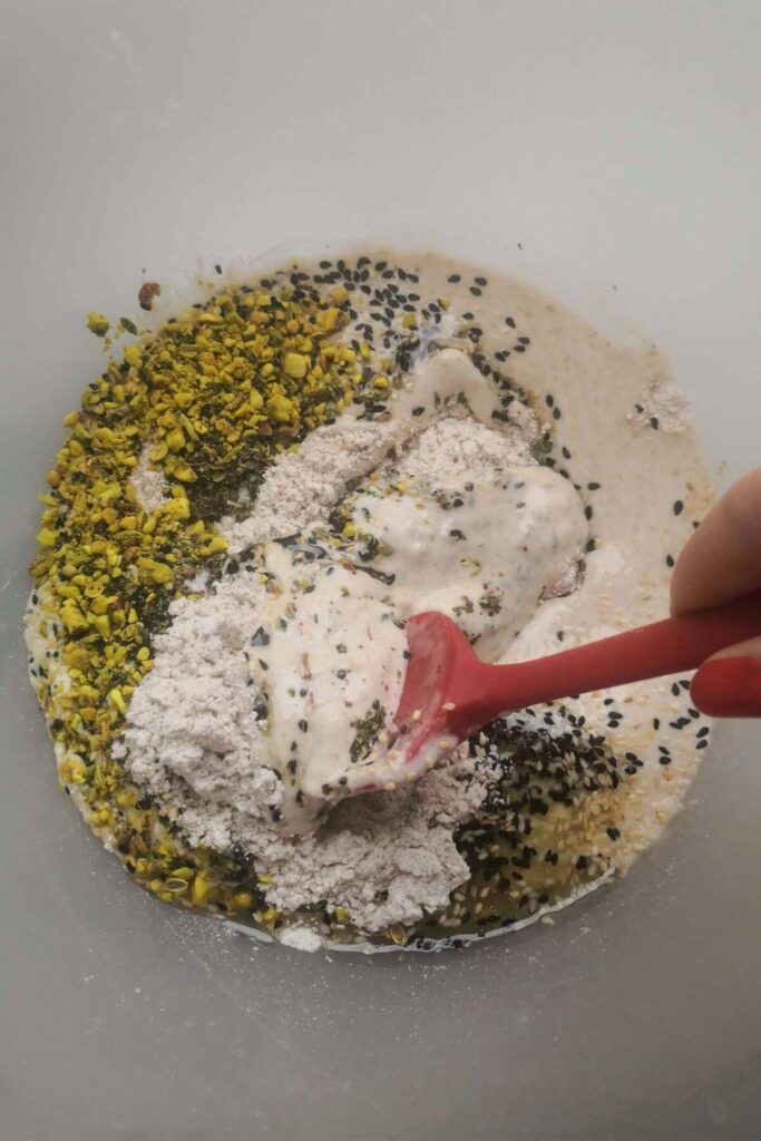 Sourdough discard ingredients in a large clear bowl with a red spatula mixing the ingredients.