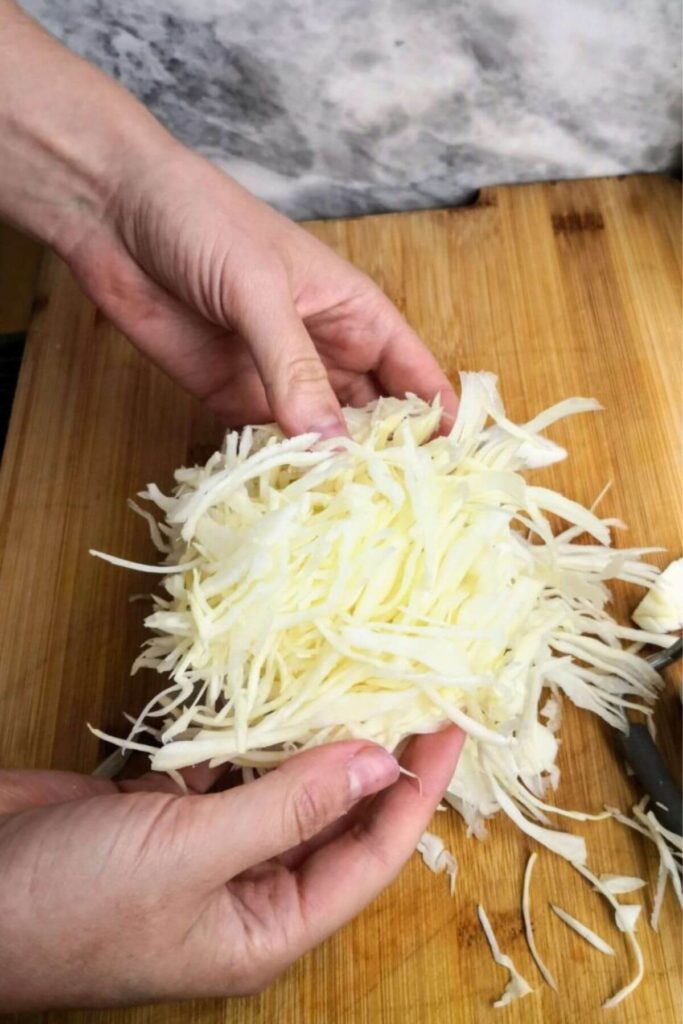 Holds holding up shredded white cabbage on a wooden board.