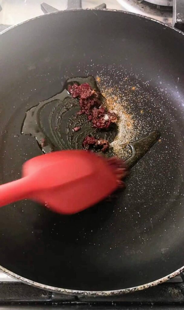 Harissa, cumin, coriander and salt being stirred in a small black pan with a red spatula.