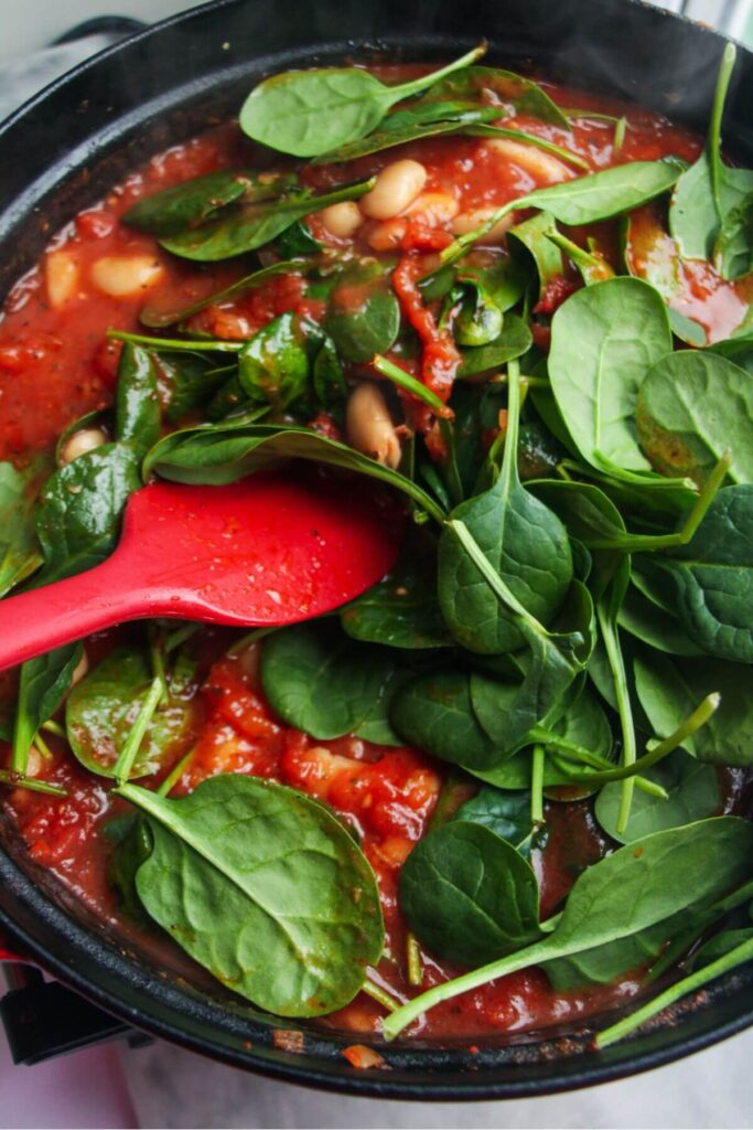 Mixing spinach leaves through white beans and tomato sauce in a large black pan.