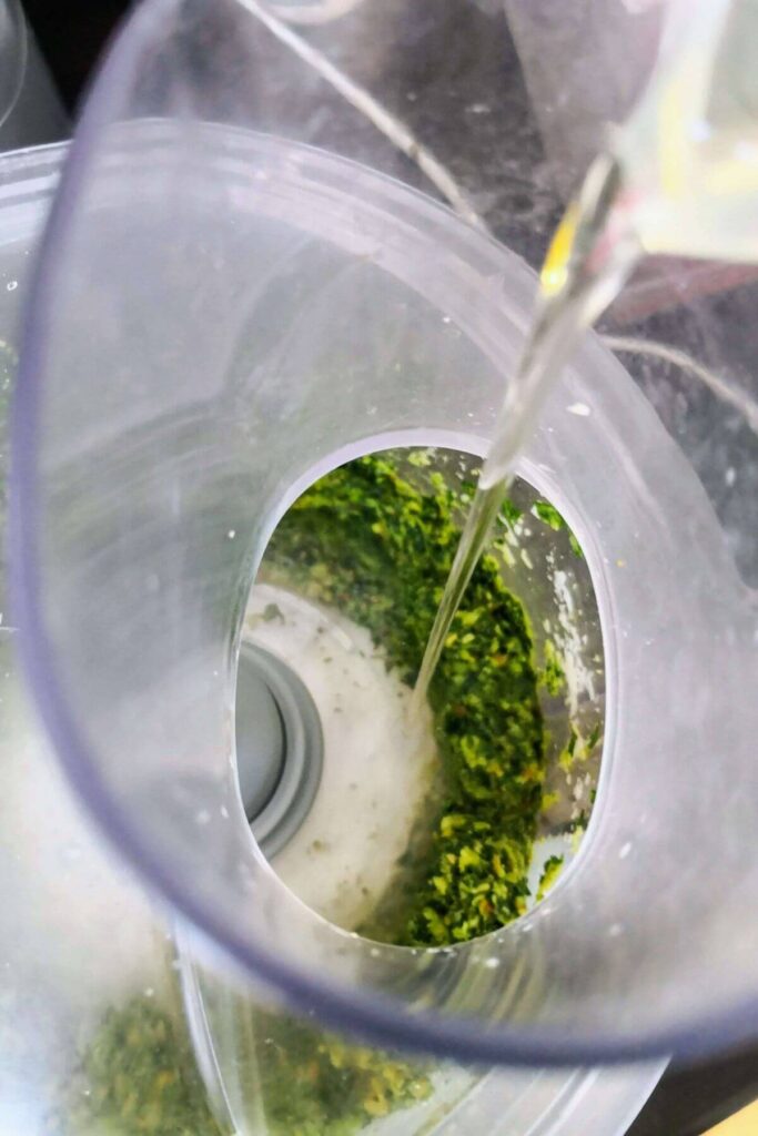 Olive oil being poured into the bowl of a food processor with pesto in it.