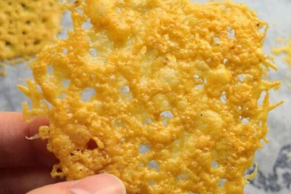 Hand holding up a parmesan crisp with more parmesan crisps on an oven tray in the background.