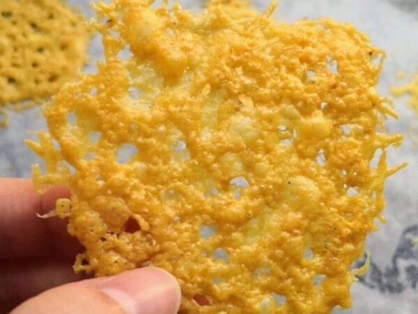 Hand holding up a parmesan crisp with more parmesan crisps on an oven tray in the background.