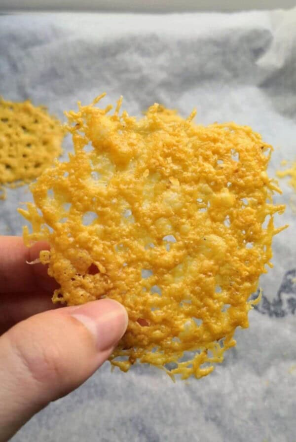 Hand holding up a parmesan crisp with more parmesan crisps on an oven tray in the background.