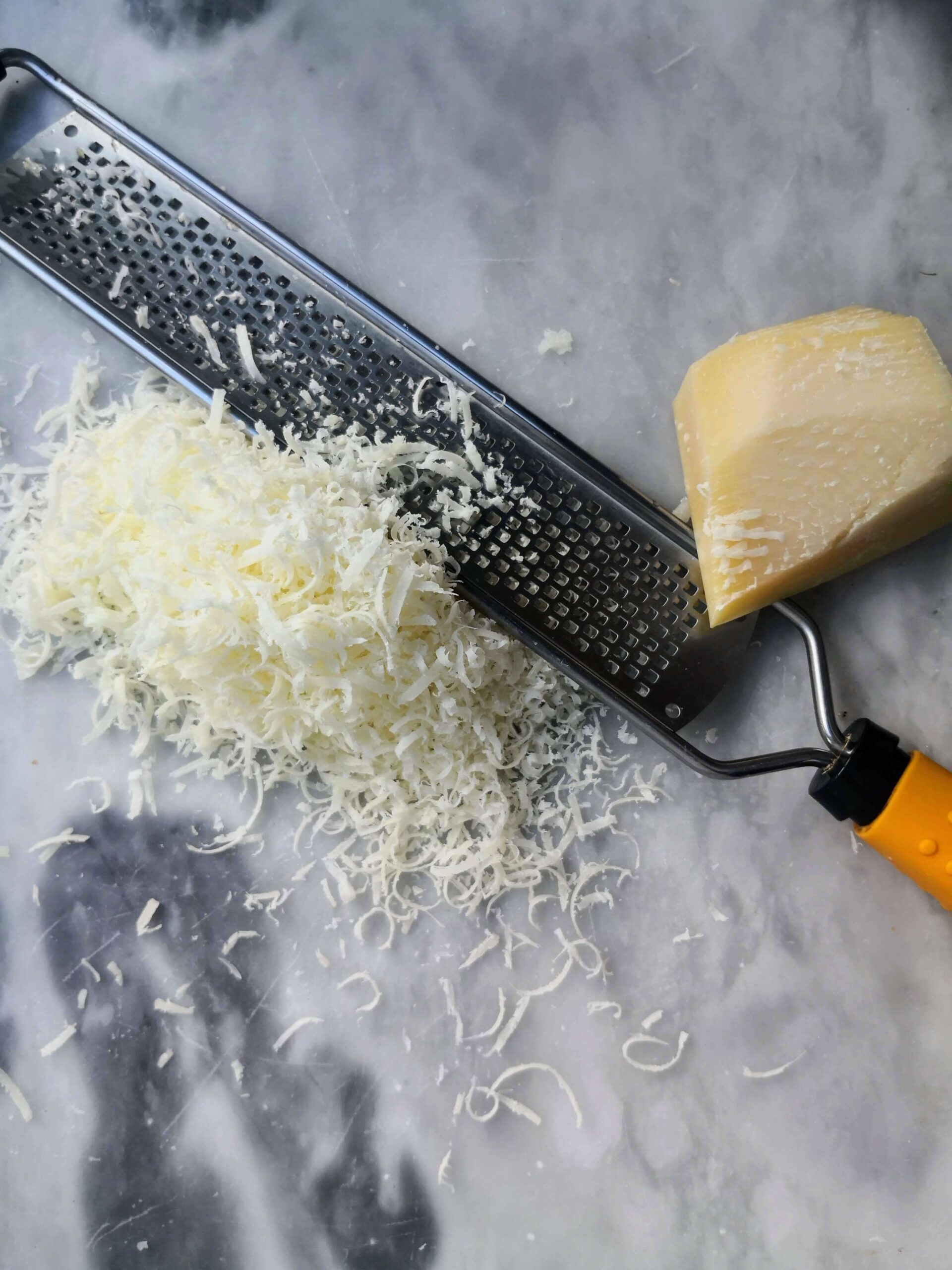 Block of parmesan cheese on a grey marble board, with a silver microplane and freshly grated parmesan next to it.
