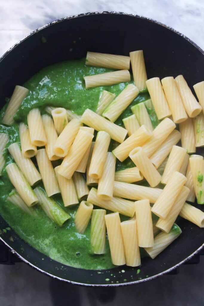 Rigatoni being added to a small black pan with green sauce in it.