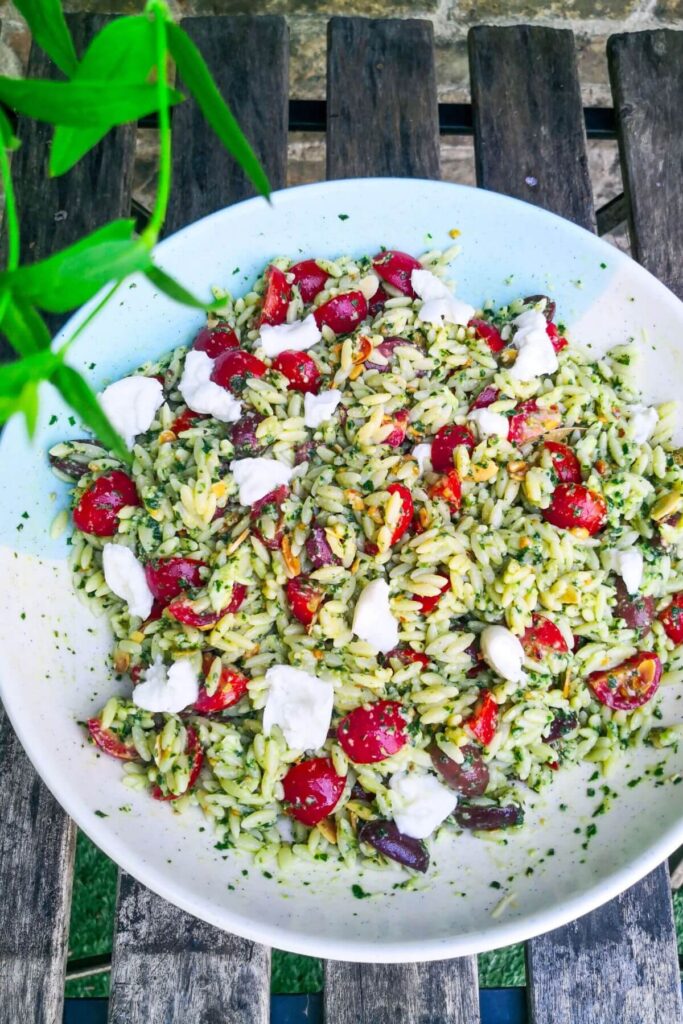 Pesto orzo salad in a large teal and white bowl on an outdoor wooden table.