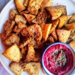 Pile of homemade pita chips on a white plate with a small bowl of beetroot hummus on the side.