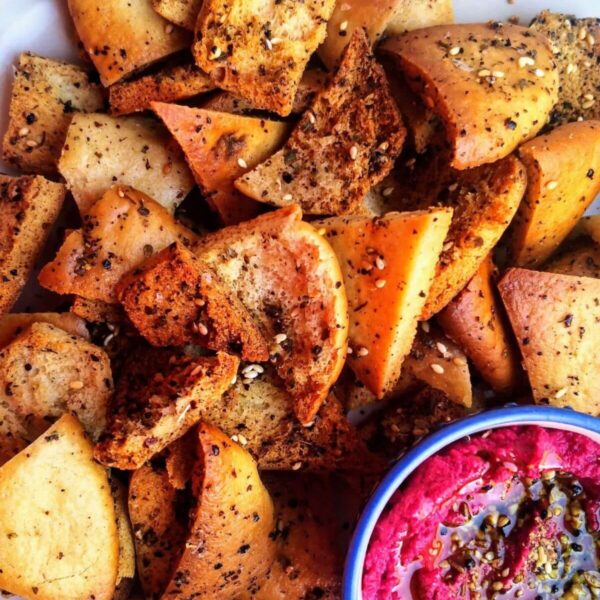 Pile of homemade pita chips on a white plate with a small bowl of beetroot hummus on the side.
