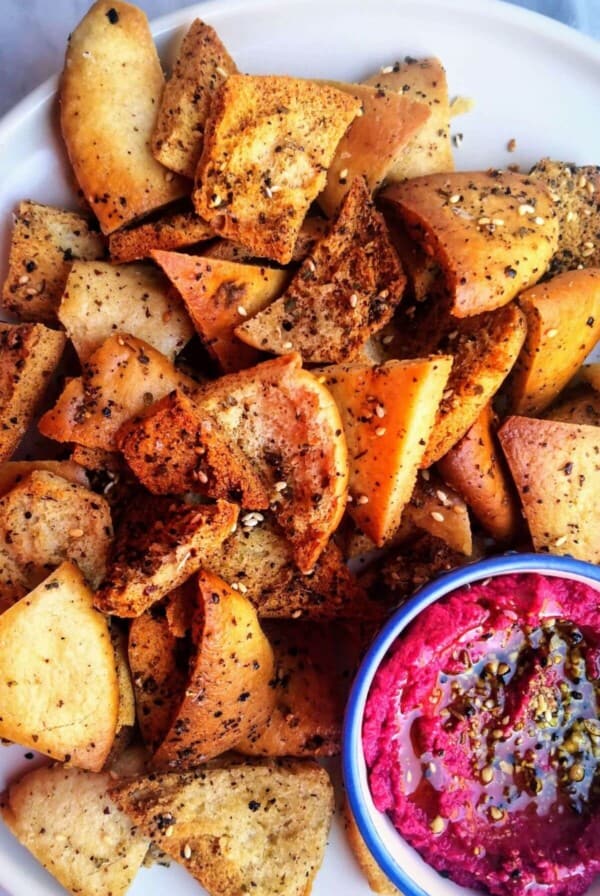 Pile of homemade pita chips on a white plate with a small bowl of beetroot hummus on the side.