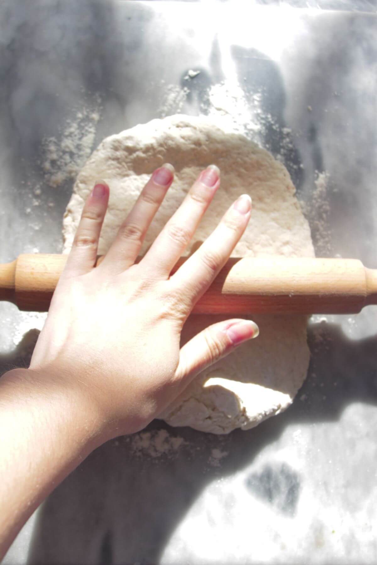 One hand using a wooden rolling pin to roll out pizza dough on a grey marble background.