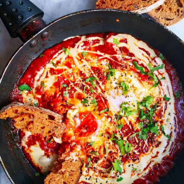 Shakshuka for one in a small black pan, on a grey marble background with sourdough placed into the sauce, and two slices of sourdough on the side.