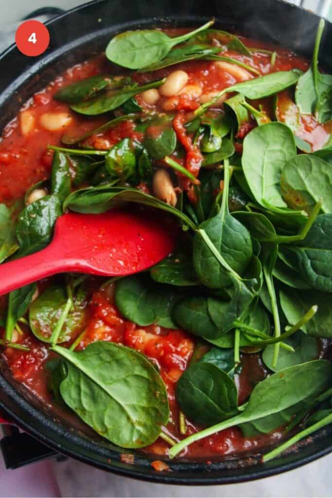 Spatula stirring spinach leaves through white beans in a tomato sauce.