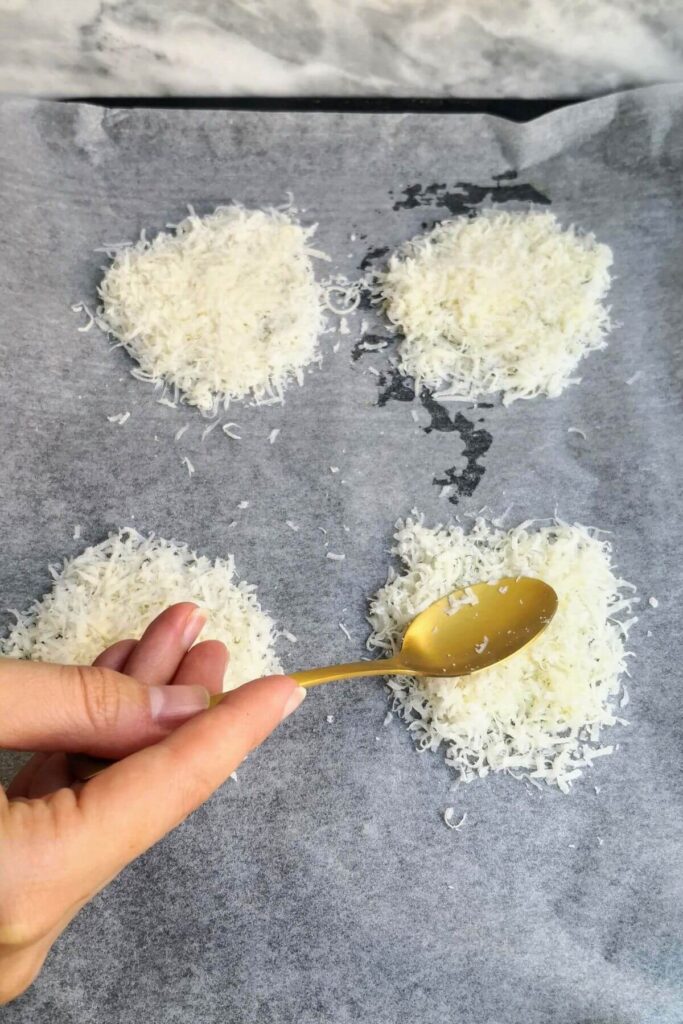 Four piles of parmesan cheese on a baking paper lined oven tray, with a hand holding a golden spoon pressing on top of one. 
