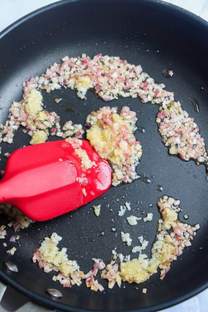 Red spatula stirring diced onion and garlic in a black pan.