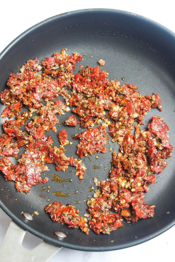 Chopped sundried tomatoes in a black pan with dried herbs.