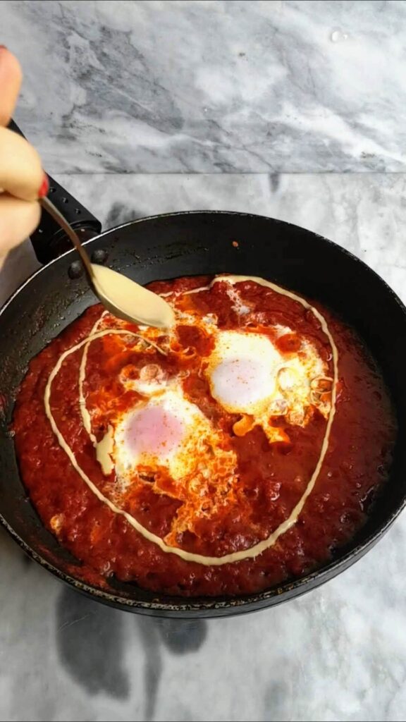 Tahini being drizzled over shakshuka in a small black pan on a grey marble background.