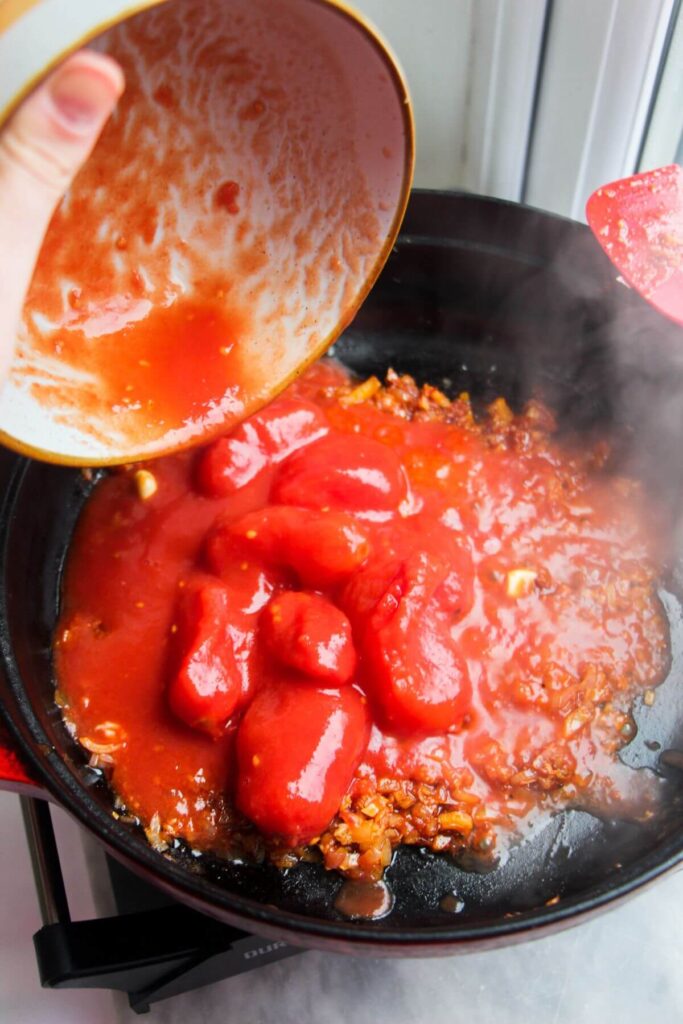 Pouring tinned tomatoes into a large black pan with shallots, nduja and garlic.